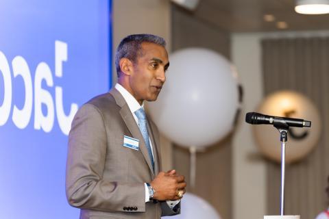 Man in beige suit, white shirt and blue tie speaking at microphone